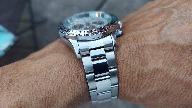 May include: A silver wristwatch with a stainless steel band is shown on a wrist. The watch has a round face with multiple dials and a rotating bezel with numbers. The band is made of rectangular links.