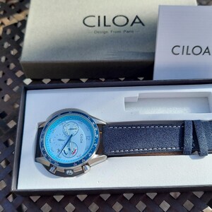 May include: A CILOA watch with a blue face and a navy blue leather strap is displayed in its box. The watch face has multiple dials and the brand name is visible. The watch is presented on a white interior.
