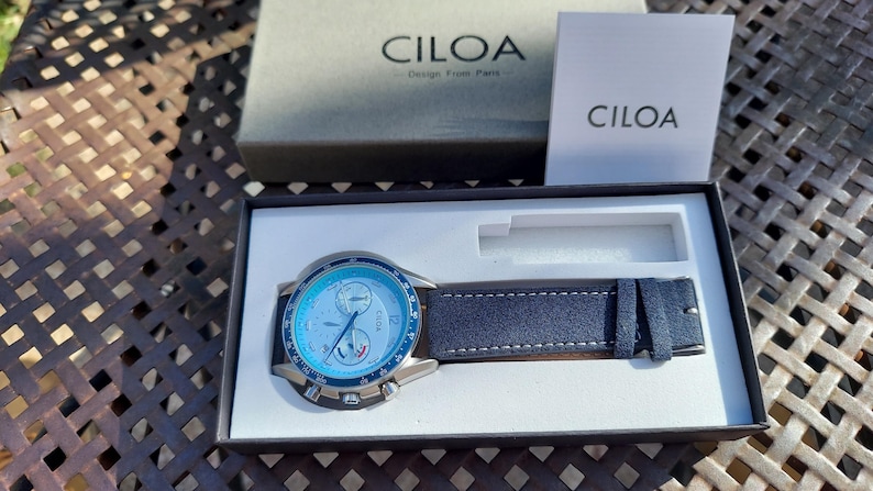 May include: A CILOA watch with a blue face and a navy blue leather strap is displayed in its box. The watch face has multiple dials and the brand name is visible. The watch is presented on a white interior.