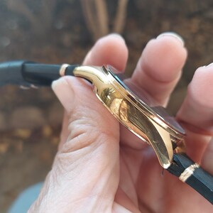 May include: A gold-toned wristwatch with a black leather band. The watch has a round face and a gold-colored bezel. The band is black with gold accents. The watch is held in a hand.