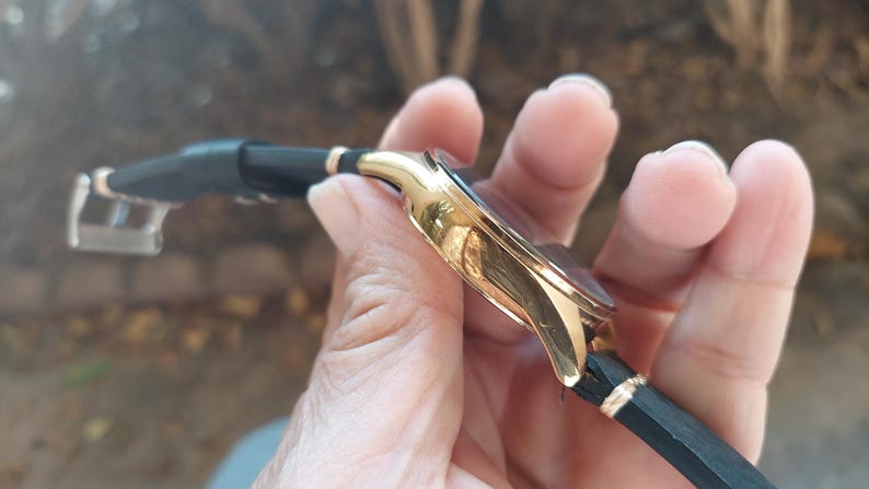 May include: A gold-toned wristwatch with a black leather band. The watch has a round face and a gold-colored bezel. The band is black with gold accents. The watch is held in a hand.