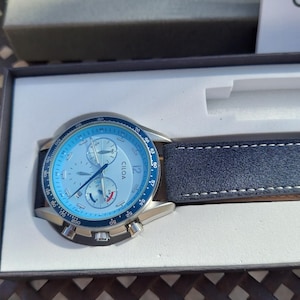 May include: A wristwatch with a light blue face and a dark blue leather band. The watch face has multiple dials and the brand name "CILOA". The watch is presented in a black box with a white interior.