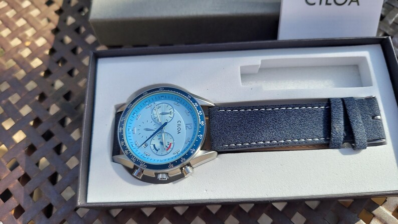May include: A wristwatch with a light blue face and a dark blue leather band. The watch face has multiple dials and the brand name "CILOA". The watch is presented in a black box with a white interior.