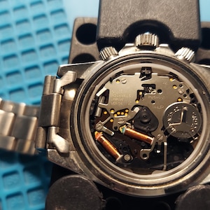 May include: A detailed view of a watch's internal workings, with the back removed. The watch features a silver metal strap and casing. The internal components, including a battery and coils, are visible. The watch is being worked on a blue surface.