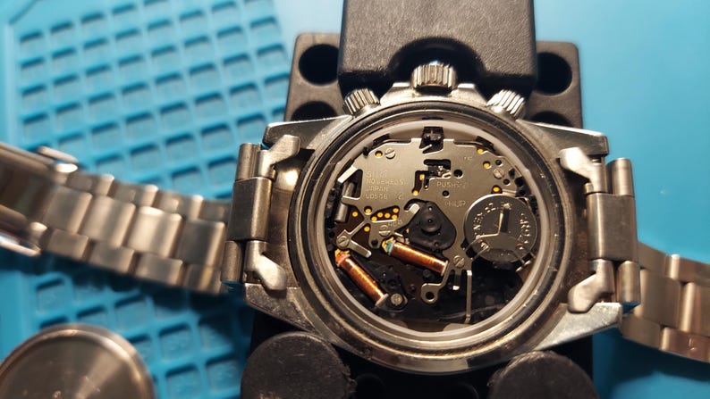 May include: A detailed view of a watch's internal workings, with the back removed. The watch features a silver metal strap and casing. The internal components, including a battery and coils, are visible. The watch is being worked on a blue surface.