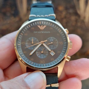 May include: A gold-toned Emporio Armani chronograph watch with a brown face and black leather strap. The watch features three sub-dials, gold hands, and the brand's logo. The watch is held in a hand.