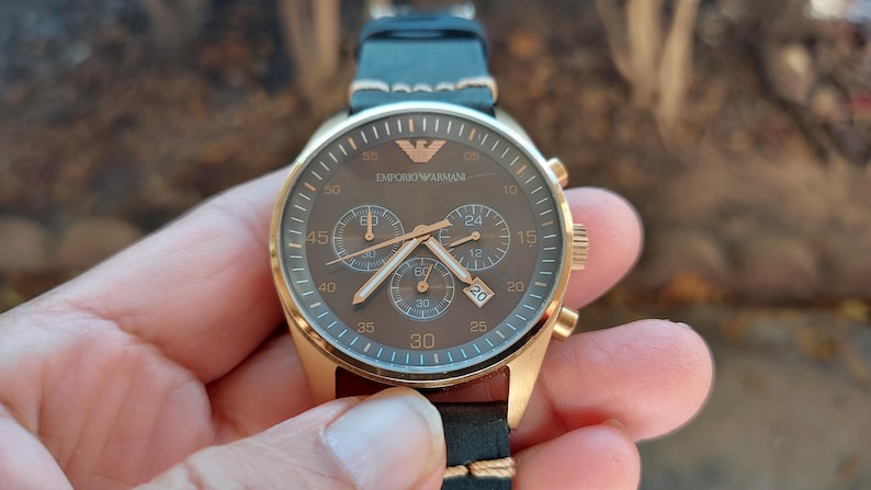May include: A gold-toned Emporio Armani chronograph watch with a brown face and black leather strap. The watch features three sub-dials, gold hands, and the brand's logo. The watch is held in a hand.