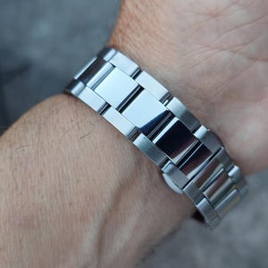 May include: A silver-toned metal watch band is fastened around a wrist. The band is composed of rectangular links with a polished, reflective surface. The watch band is a classic style, suitable for everyday wear.