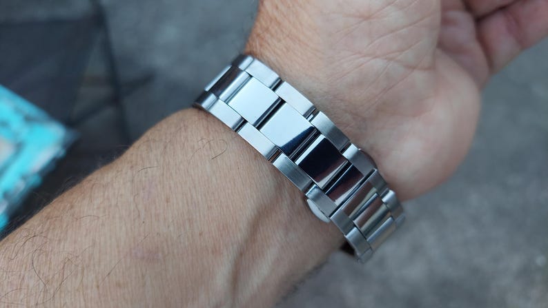 May include: A silver-toned metal watch band is fastened around a wrist. The band is composed of rectangular links with a polished, reflective surface. The watch band is a classic style, suitable for everyday wear.