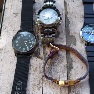 May include: An assortment of wristwatches with various band styles and face designs. One watch has a black nylon band and a dark face with white numerals. Another has a silver metal band and a light-colored face. A third watch has a blue face and a black rubber band.