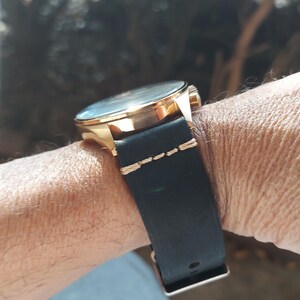 May include: A gold-toned wristwatch with a black leather band is shown on a wrist. The watch face is visible, and the band is secured with a buckle. The band is stitched with gold thread.