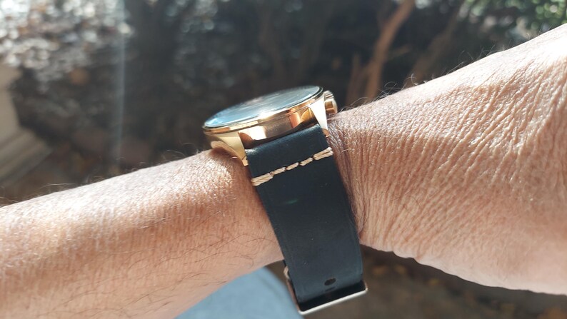 May include: A gold-toned wristwatch with a black leather band is shown on a wrist. The watch face is visible, and the band is secured with a buckle. The band is stitched with gold thread.