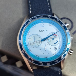 May include: A CILOA chronograph watch with a white face, blue accents, and a navy blue leather strap. The watch features three sub-dials and a date display. The watch is set against a neutral background.