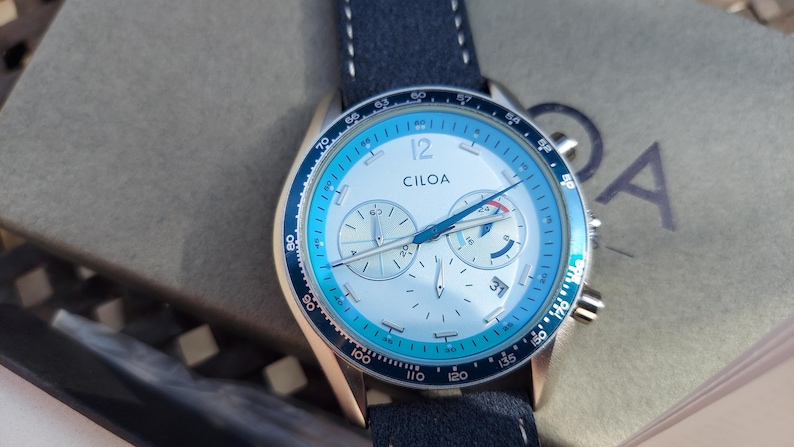 May include: A CILOA chronograph watch with a white face, blue accents, and a navy blue leather strap. The watch features three sub-dials and a date display. The watch is set against a neutral background.