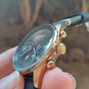 May include: Gold-toned wristwatch with a dark gray face and black leather band. The watch features three sub-dials and a gold crown. The brand name "Armani" is visible on the watch face. The watch is a stylish accessory.