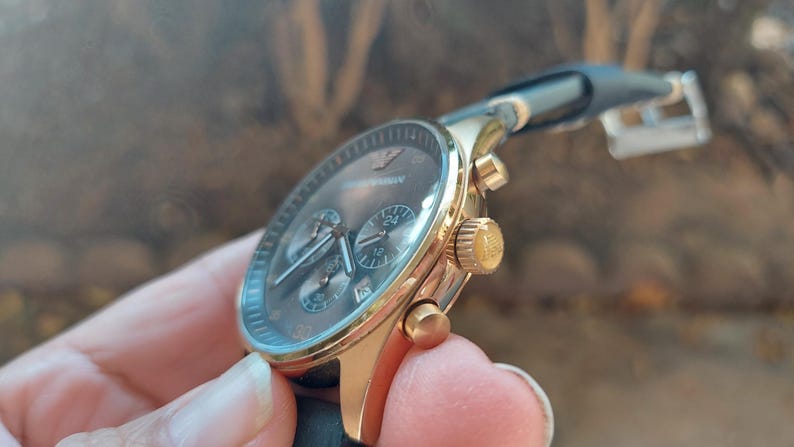 May include: Gold-toned wristwatch with a dark gray face and black leather band. The watch features three sub-dials and a gold crown. The brand name "Armani" is visible on the watch face. The watch is a stylish accessory.