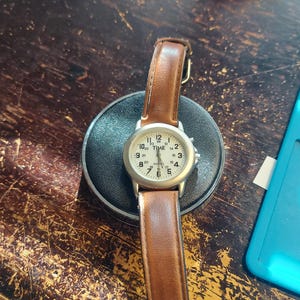 May include: A wristwatch with a silver-colored case and a light-colored face, featuring black numerals and hands. The watch has a brown leather band. The watch is resting on a black circular base.