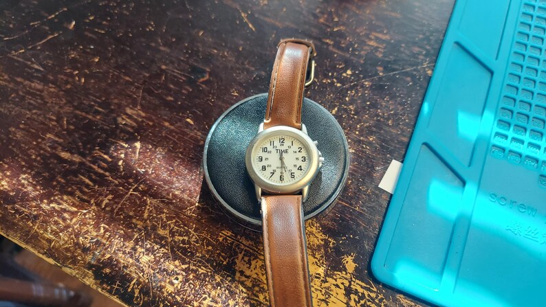 May include: A wristwatch with a silver-colored case and a light-colored face, featuring black numerals and hands. The watch has a brown leather band. The watch is resting on a black circular base.