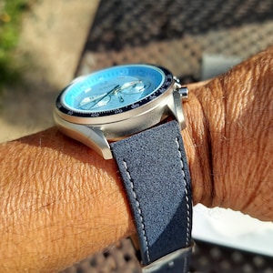 May include: A silver wristwatch with a light blue face and a dark blue suede strap is shown on a person's wrist. The watch has a black bezel and silver hands. The strap is stitched with white thread.