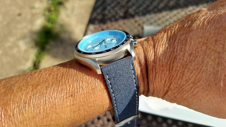 May include: A silver wristwatch with a light blue face and a dark blue suede strap is shown on a person's wrist. The watch has a black bezel and silver hands. The strap is stitched with white thread.