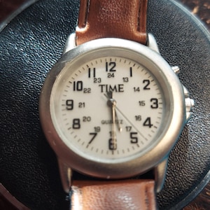 May include: A silver-toned wristwatch with a white face and brown leather band. The watch face displays the word "TIME" above the hands, with black numerals marking the hours and minutes. The watch is a quartz movement.