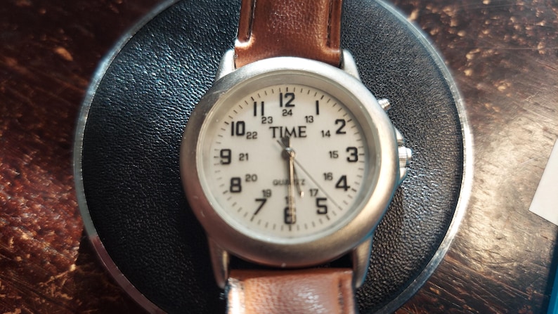May include: A silver-toned wristwatch with a white face and brown leather band. The watch face displays the word "TIME" above the hands, with black numerals marking the hours and minutes. The watch is a quartz movement.