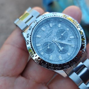 May include: A silver wristwatch with a textured gray face and three sub-dials. The watch has a silver band and a rotating bezel with black markings. The watch is held in a hand.
