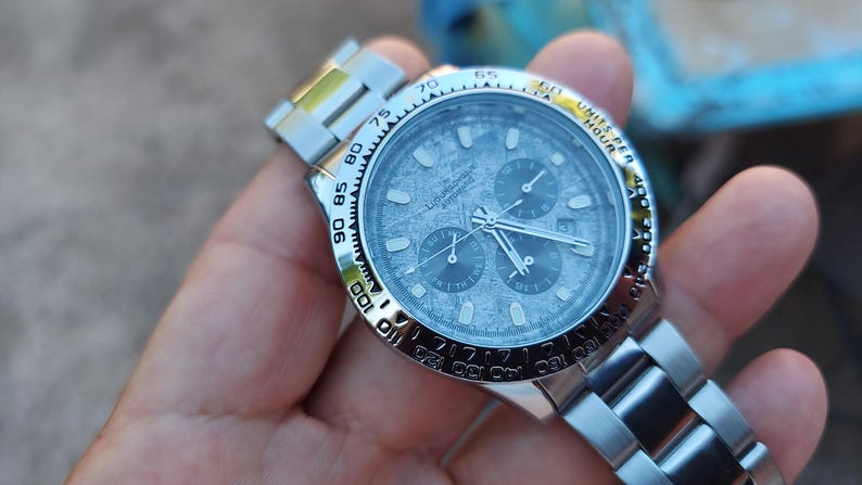 May include: A silver wristwatch with a textured gray face and three sub-dials. The watch has a silver band and a rotating bezel with black markings. The watch is held in a hand.