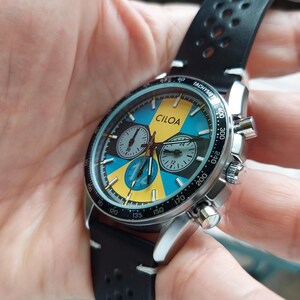 Pu&ograve; includere: Un orologio cronografo CILOA con cassa in acciaio inossidabile color argento e cinturino in pelle nera traforata. Il quadrante presenta un vivace motivo geometrico giallo e blu, tre sotto-quadranti e una lunetta tachimetrica nera con segni bianchi.