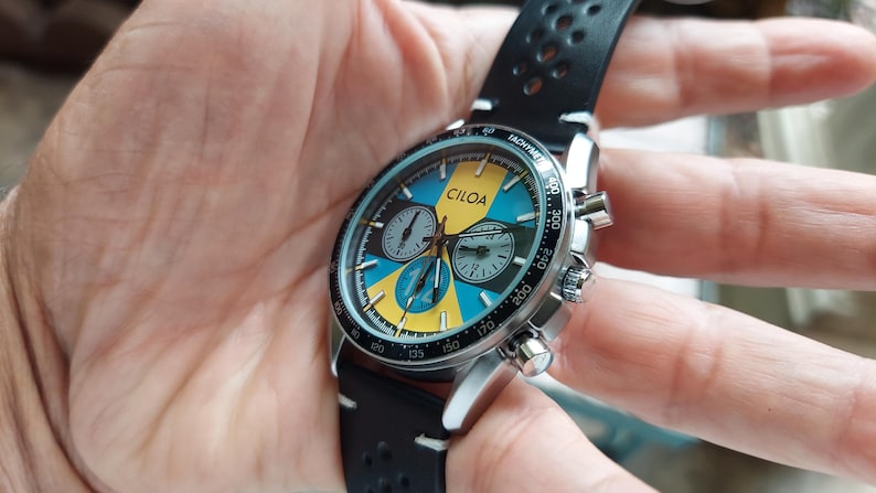 Pu&ograve; includere: Un orologio cronografo CILOA con cassa in acciaio inossidabile color argento e cinturino in pelle nera traforata. Il quadrante presenta un vivace motivo geometrico giallo e blu, tre sotto-quadranti e una lunetta tachimetrica nera con segni bianchi.