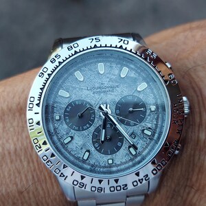 May include: A silver wristwatch with a textured gray face and three sub-dials. The watch has a silver bezel with black markings and a silver band. The watch face has the text "LUXURIOUS AUTOMATIC".