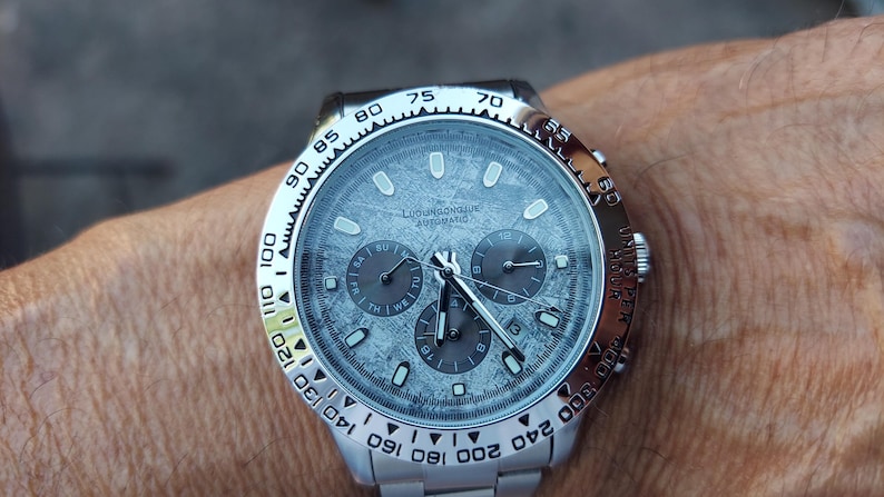 May include: A silver wristwatch with a textured gray face and three sub-dials. The watch has a silver bezel with black markings and a silver band. The watch face has the text "LUXURIOUS AUTOMATIC".