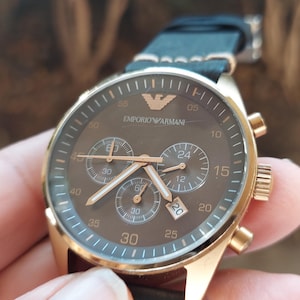 May include: A gold-toned Emporio Armani chronograph watch with a brown face and black leather strap. The watch features three sub-dials, gold hands, and the brand's logo. The watch case is approximately 40mm in diameter.