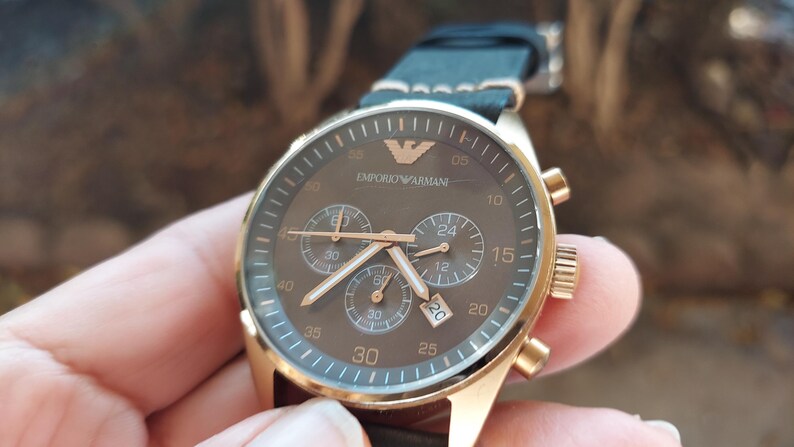 May include: A gold-toned Emporio Armani chronograph watch with a brown face and black leather strap. The watch features three sub-dials, gold hands, and the brand's logo. The watch case is approximately 40mm in diameter.