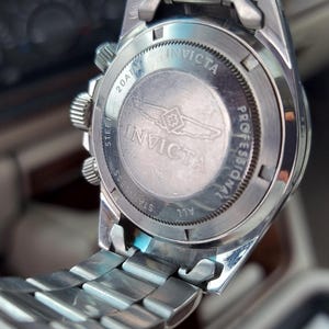 May include: Close-up of the back of a silver Invicta Professional watch. The watch has a stainless steel strap and a round face with the Invicta logo and text. The watch is set against a blurred background.