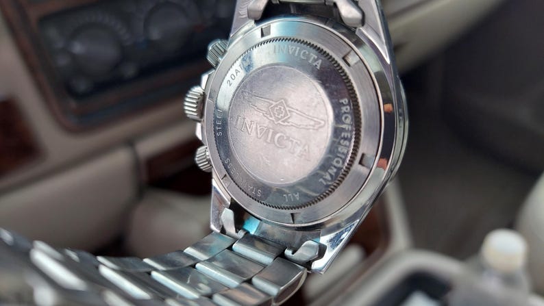 May include: Close-up of the back of a silver Invicta Professional watch. The watch has a stainless steel strap and a round face with the Invicta logo and text. The watch is set against a blurred background.