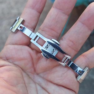 May include: A silver-toned watch clasp, featuring a butterfly deployment design. The clasp is made of polished metal and has a rectangular shape with a series of interlocking segments. The watch clasp is designed to secure a watch band.