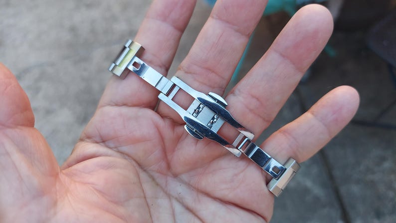 May include: A silver-toned watch clasp, featuring a butterfly deployment design. The clasp is made of polished metal and has a rectangular shape with a series of interlocking segments. The watch clasp is designed to secure a watch band.