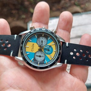 Pu&ograve; includere: Un orologio cronografo CILOA con cassa rotonda color argento e cinturino in pelle nera traforata. Il quadrante presenta un vivace motivo geometrico blu e giallo con tre sotto-quadranti, indici bianchi e una lunetta tachimetrica nera.