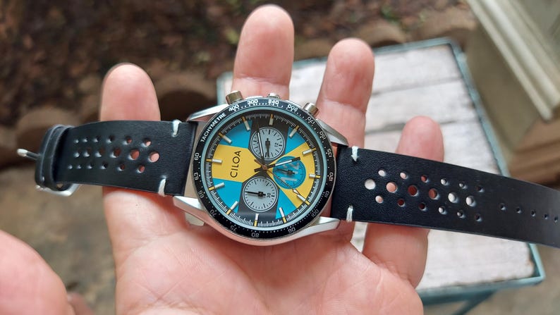 Pu&ograve; includere: Un orologio cronografo CILOA con cassa rotonda color argento e cinturino in pelle nera traforata. Il quadrante presenta un vivace motivo geometrico blu e giallo con tre sotto-quadranti, indici bianchi e una lunetta tachimetrica nera.