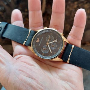 May include: A gold-toned wristwatch with a dark brown face and black leather band. The watch features three sub-dials and a date display. The black leather band has gold stitching and a buckle closure.
