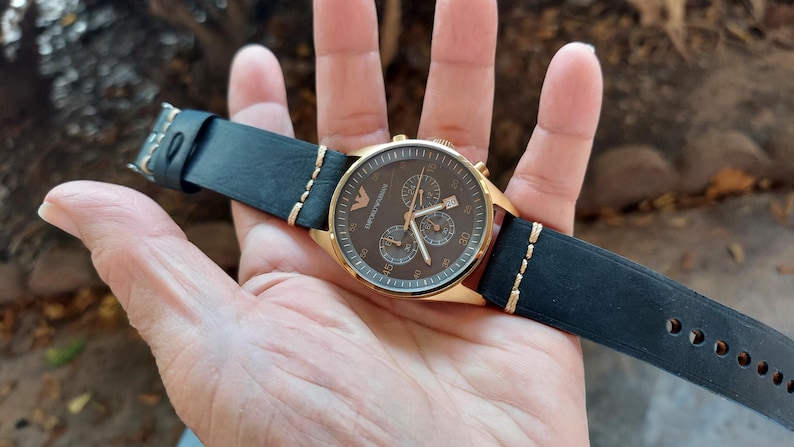 May include: A gold-toned wristwatch with a dark brown face and black leather band. The watch features three sub-dials and a date display. The black leather band has gold stitching and a buckle closure.