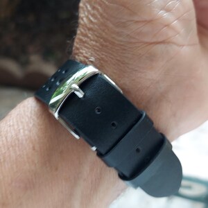 Pu&ograve; includere: Un cinturino per orologio nero con finitura opaca liscia e una fibbia in metallo color argento lucido. Il cinturino include due passanti fissi e una serie di piccoli fori circolari per la regolazione, fissati con una chiusura a fibbia standard.