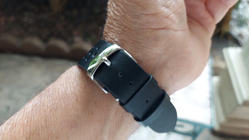 Pu&ograve; includere: Un cinturino per orologio nero con finitura opaca liscia e una fibbia in metallo color argento lucido. Il cinturino include due passanti fissi e una serie di piccoli fori circolari per la regolazione, fissati con una chiusura a fibbia standard.