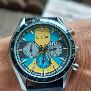 Pu&ograve; includere: Un orologio cronografo color argento con una lunetta nera dotata di scala tachimetrica. Il quadrante presenta un motivo geometrico giallo e blu, il marchio CILOA, tre sotto-quadranti e il numero 112 stampato in blu sul sotto-quadrante inferiore.