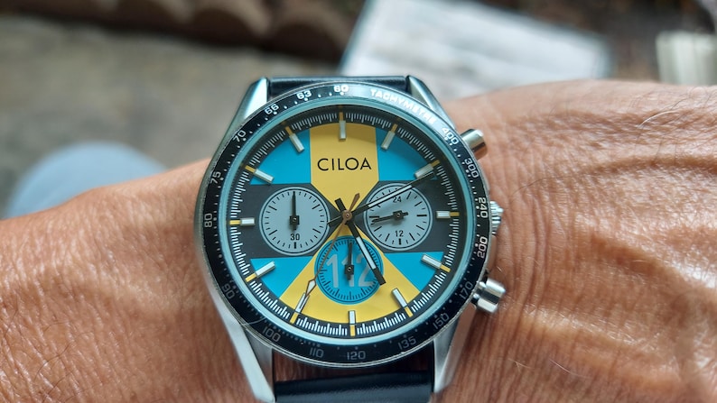 Pu&ograve; includere: Un orologio cronografo color argento con una lunetta nera dotata di scala tachimetrica. Il quadrante presenta un motivo geometrico giallo e blu, il marchio CILOA, tre sotto-quadranti e il numero 112 stampato in blu sul sotto-quadrante inferiore.