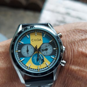 Pu&ograve; includere: Un orologio cronografo color argento con quadrante rotondo caratterizzato da un motivo geometrico giallo e blu. Il quadrante riporta il marchio CILOA, tre sotto-quadranti e una scala tachimetrica sulla lunetta esterna. Cinturino nero.