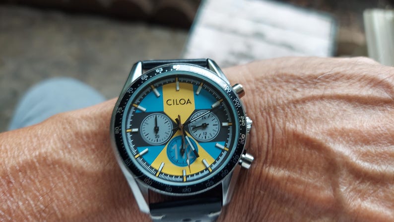 Pu&ograve; includere: Un orologio cronografo color argento con quadrante rotondo caratterizzato da un motivo geometrico giallo e blu. Il quadrante riporta il marchio CILOA, tre sotto-quadranti e una scala tachimetrica sulla lunetta esterna. Cinturino nero.
