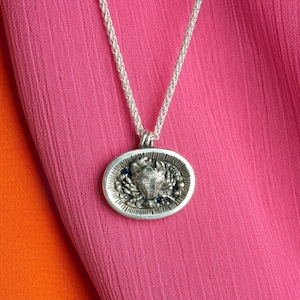 Pode incluir: Um colar de corrente de prata com um pingente oval de prata que apresenta um design de caranguejo detalhado com acentos azuis.