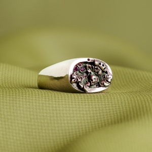 May include: A silver ring with an oval-shaped design featuring a skull surrounded by intricate details and red gemstones.